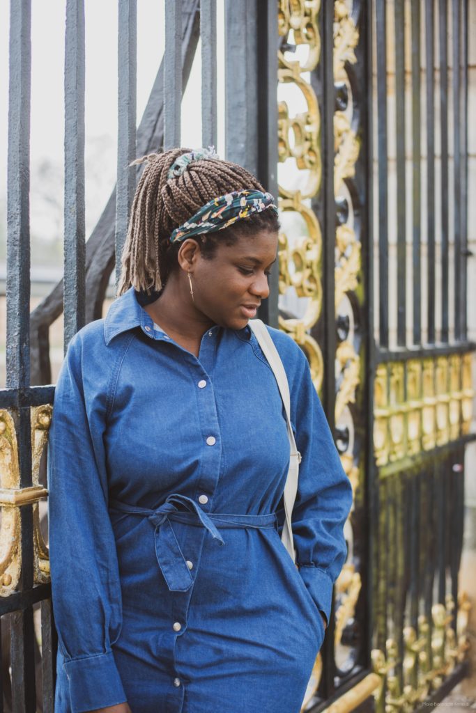 Adelana is standing by a gate black and gold wearing a denim blue dress. She's looking down