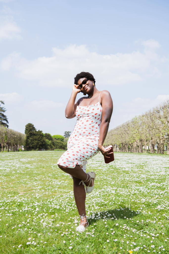 Adelana porte une robe avec des motifs bisous avec une pochette rouge en pinatex. Elle est dans le milieu d'un parc avec des marguerites. 