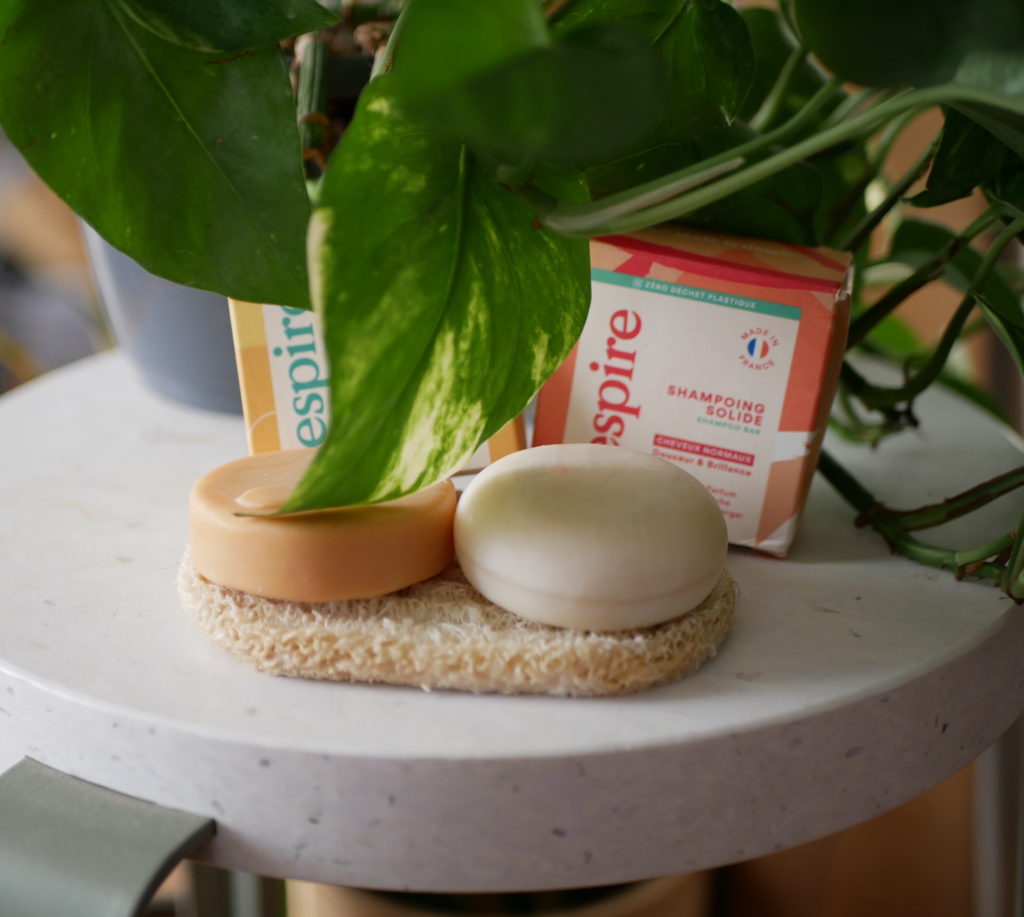 Les produits shampoing et après shampoing de Respire posé sur un porte savon en loofah. Une plante verte en arrière plan donc une feuille qui tombe sur l'après shampoing solide